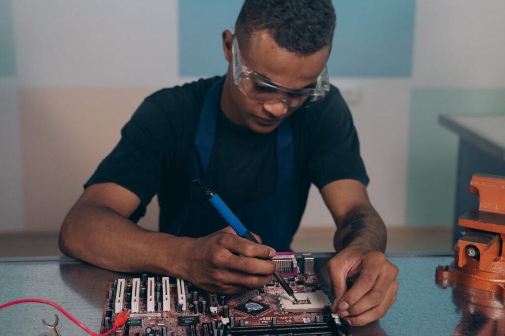 A technician wearing safety glasses works intently on a computer motherboard with a soldering iron.